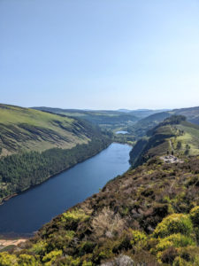 A view of the Glendalough upper lake from the top of the hike. The sky is clear and blue and reflected on the water. The hills are green.