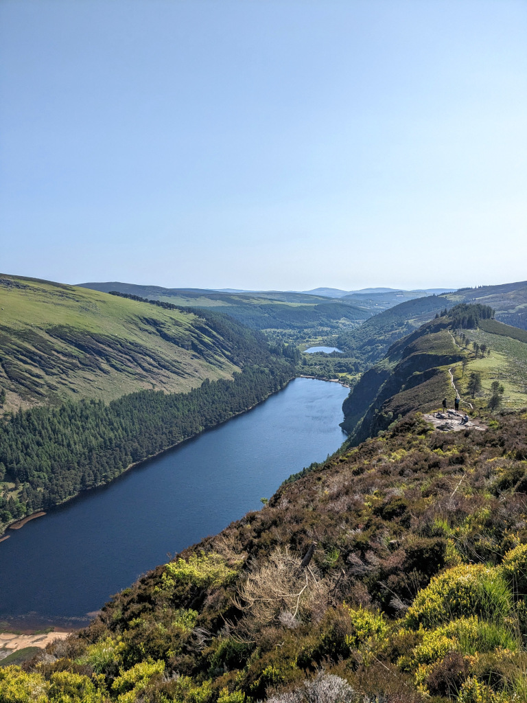 A view of the Glendalough upper lake from the top of the hike. The sky is clear and blue and reflected on the water. The hills are green.